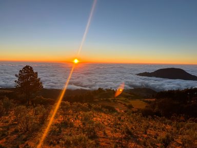 Atardecer sobre un mar de nubes, con un sol resplandeciente y montañas al fondo.